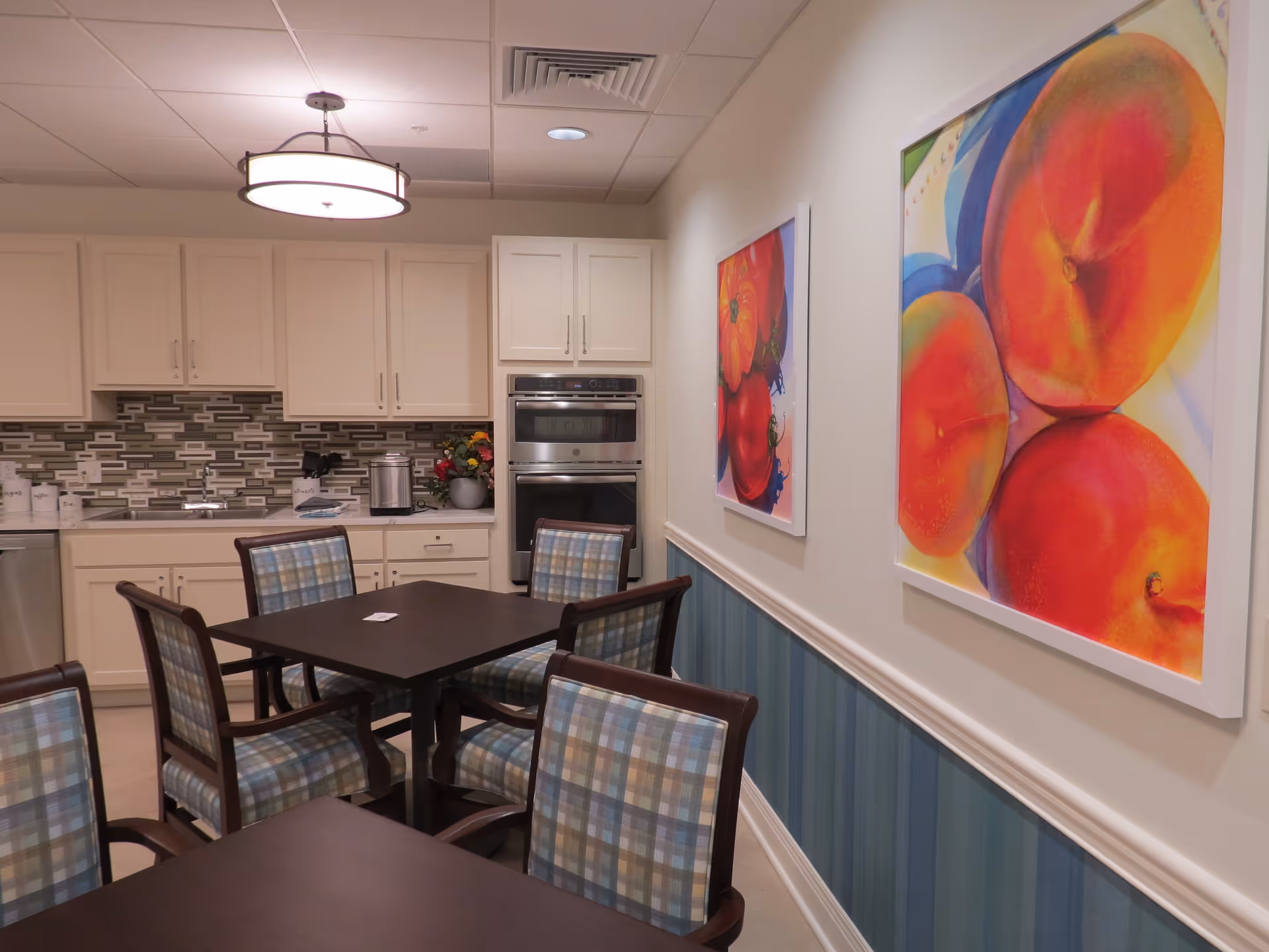 A cozy kitchen and dining area with white cabinets, a tiled backsplash, and stainless steel appliances. There are two dark wood tables with plaid upholstered chairs. The walls are decorated with colorful framed paintings of fruit.