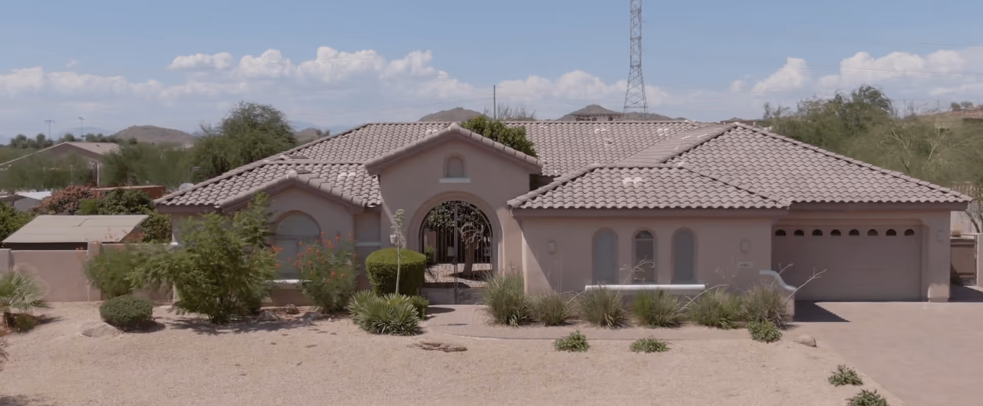 Single-story residential building with a tiled roof, arched entrance gate, and desert landscaping including bushes and small plants under a partly cloudy sky.