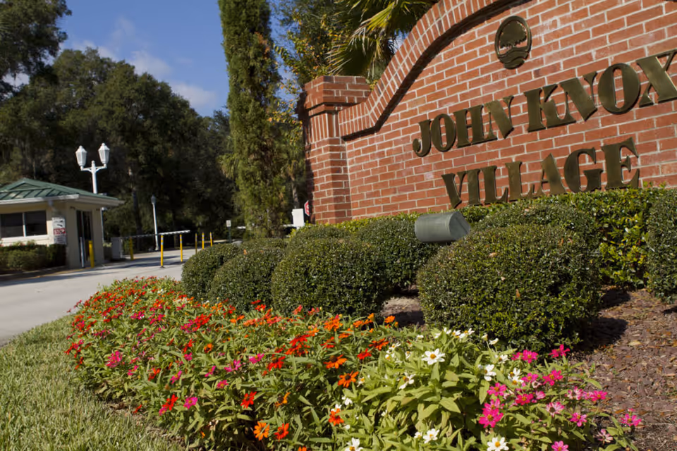 Entrance to John Knox Village of Central Florida featuring a brick sign with the facility name, neatly trimmed bushes, colorful flowers, and a security gatehouse in the background under a partly cloudy sky.