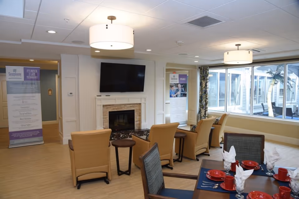 A cozy common area in a senior living facility featuring a fireplace with a mounted flat-screen TV above it, surrounded by beige armchairs and small tables. To the right, there is a dining table set with red plates, cups, and white folded napkins. Large windows let in natural light, and informational banners are visible in the background.