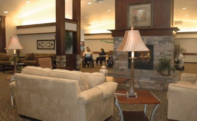 Spacious assisted-living communal lounge with beige sofas, side tables and lamps, a central stone fireplace, and residents in wheelchairs in the background.