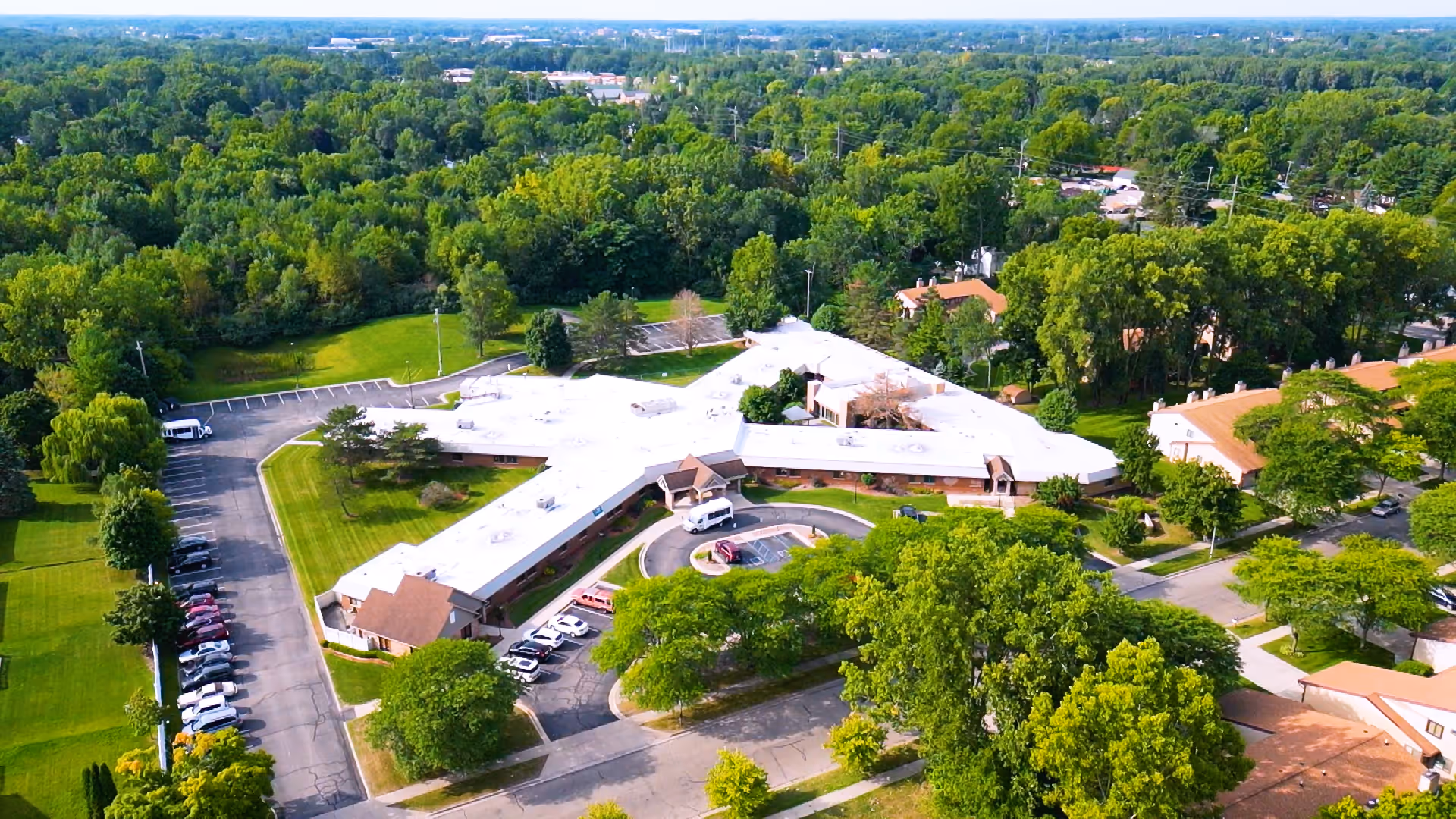Aerial view of Medilodge of Capital Area facility surrounded by lush green trees and lawns, with parking lots and roads visible around the building.