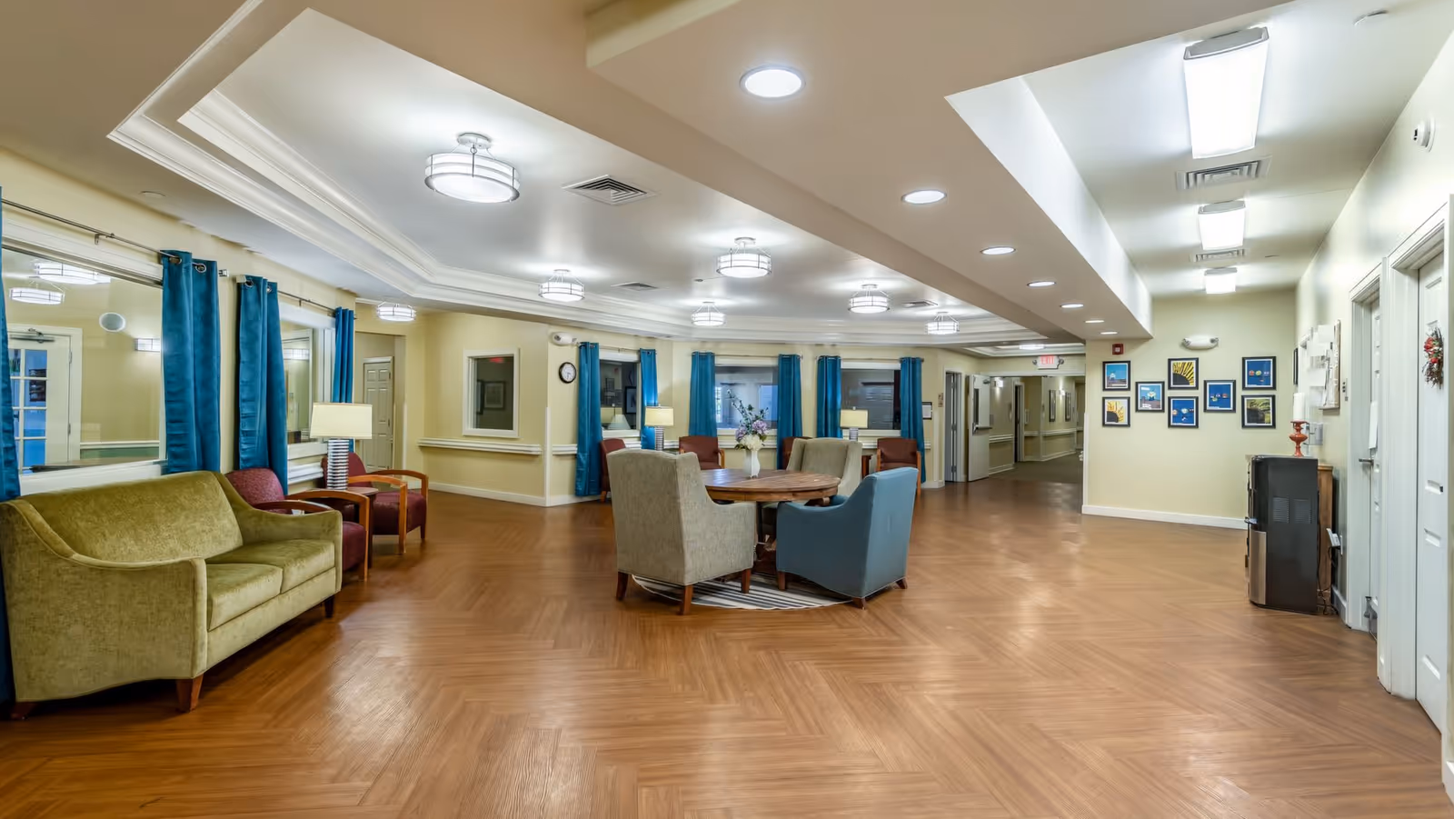 A spacious, well-lit common area in a senior living facility with wooden flooring, a round table surrounded by four upholstered chairs, a green sofa, and several armchairs. The walls are light-colored with blue curtains on the windows, framed artwork, and multiple ceiling lights. There are hallways leading to other rooms in the background.