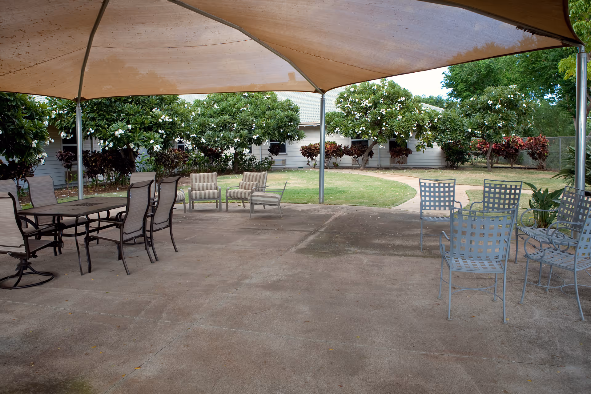 Shaded outdoor patio with metal and cushioned chairs and tables facing a grassy courtyard and low building.