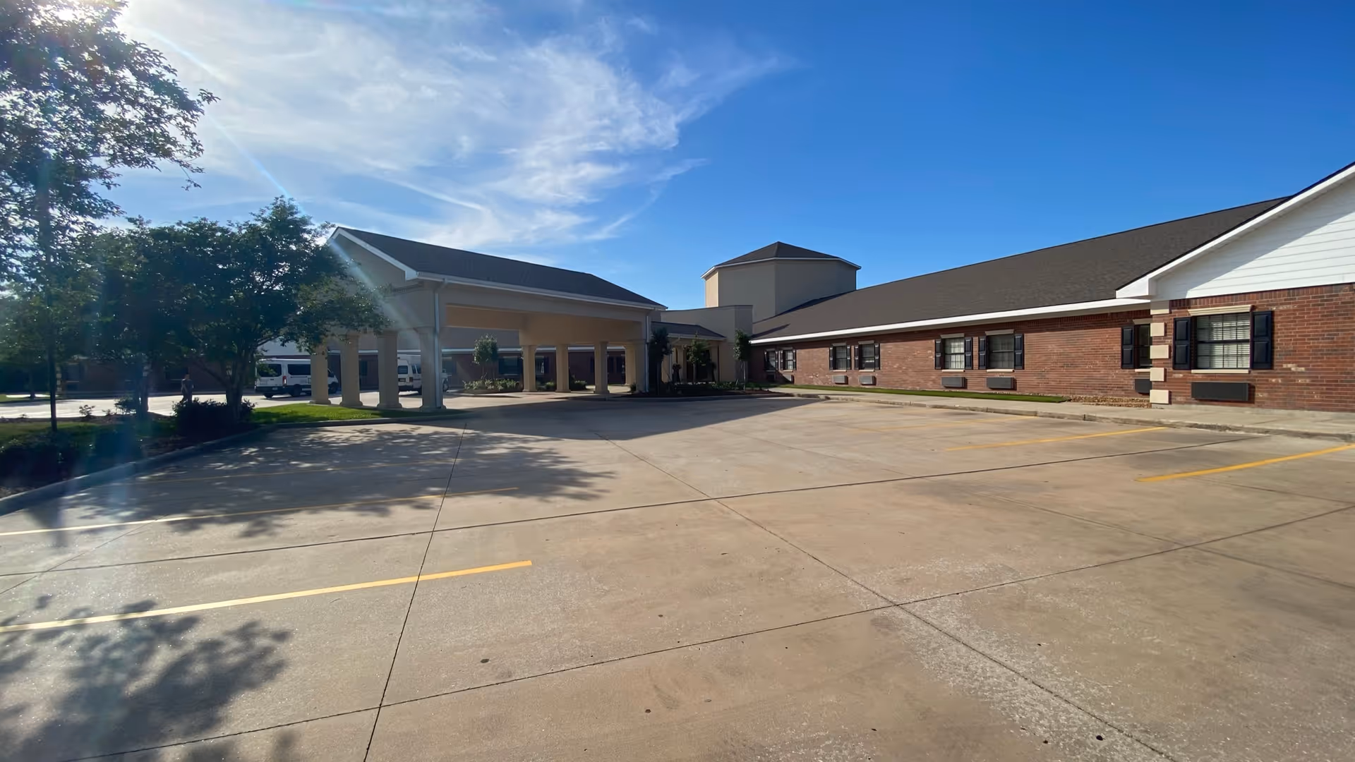 Exterior view of a senior living facility building with a covered entrance, brick walls, multiple windows, and a large empty parking lot under a clear blue sky.
