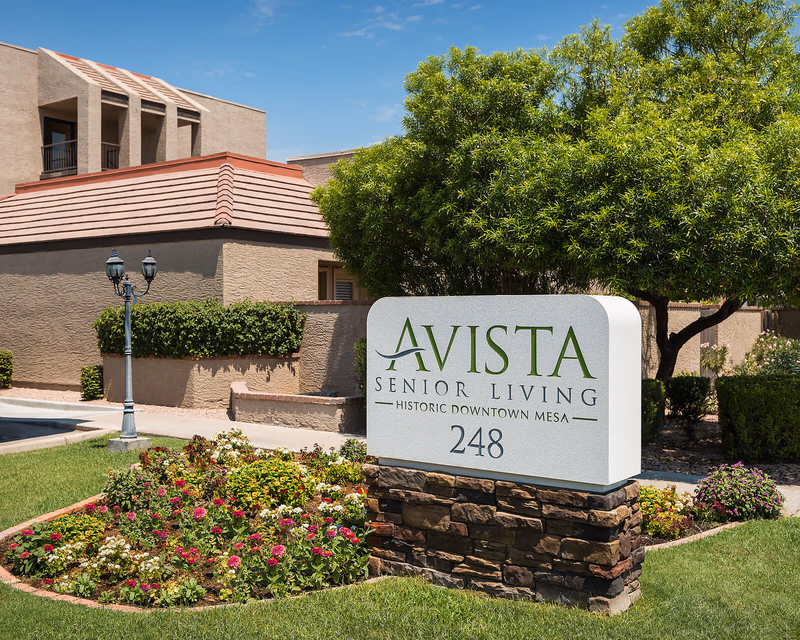 Entrance sign reading 'AVISTA Senior Living Historic Downtown Mesa 248' on a stone base surrounded by flowers and trees in front of a beige building.