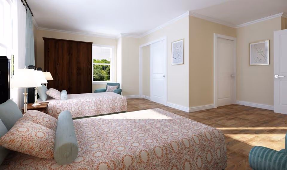 A bright bedroom with two beds covered in patterned bedspreads, a wooden wardrobe, a teal armchair, two lamps on bedside tables, and two framed abstract artworks on the walls. Sunlight streams in through a window with light curtains.