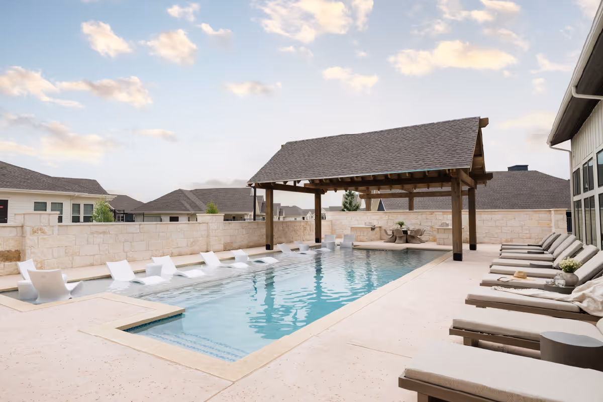 Outdoor swimming pool area with lounge chairs lined up on the right side and white poolside chairs partially submerged in the water. A wooden pergola with a shingled roof is situated at the far end of the pool. The area is enclosed by a stone wall, and residential-style buildings are visible in the background under a partly cloudy sky.