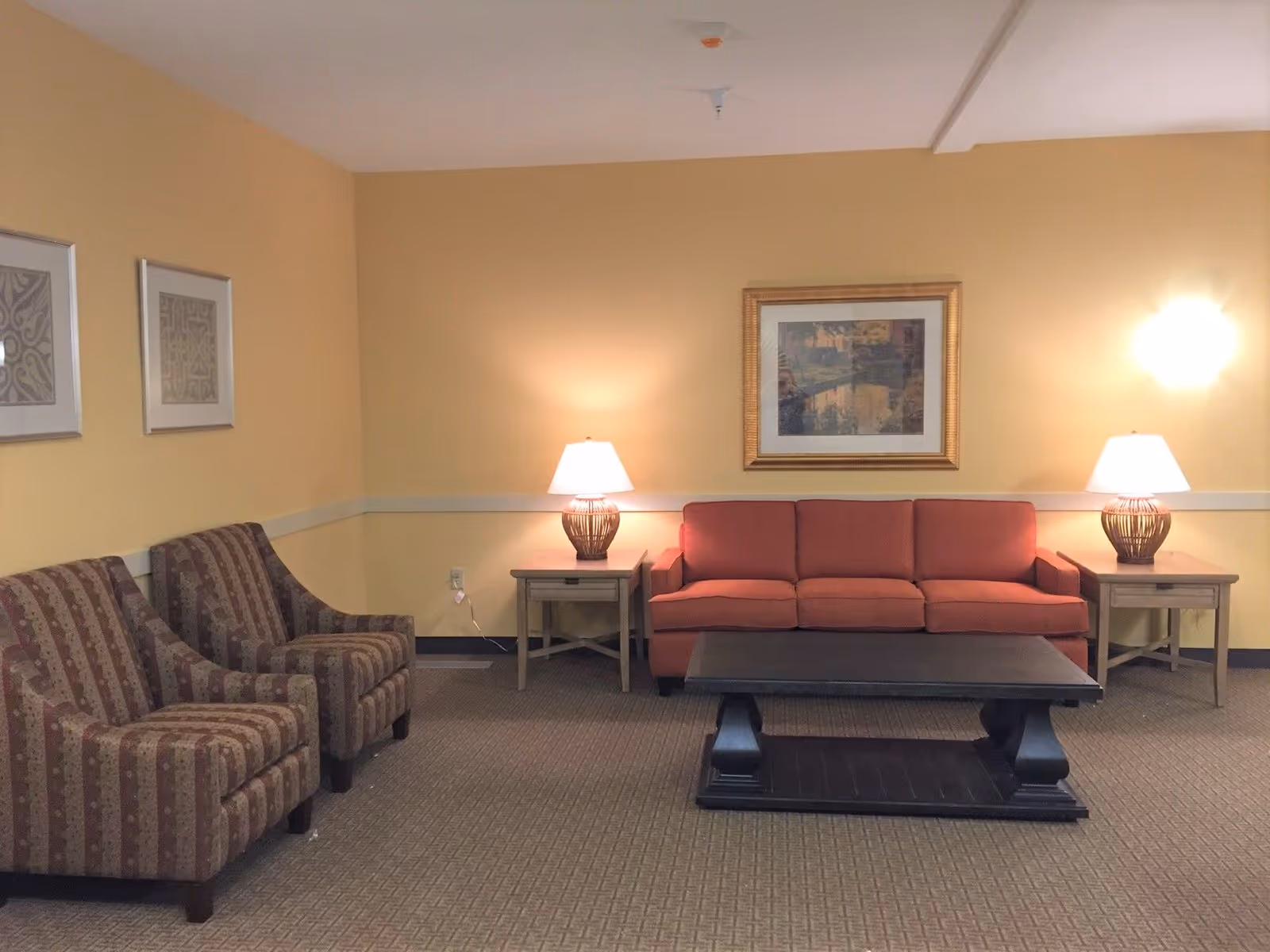 A cozy living room with a red sofa, two patterned armchairs, a dark wooden coffee table, two side tables with lamps, and framed artwork on the yellow walls.
