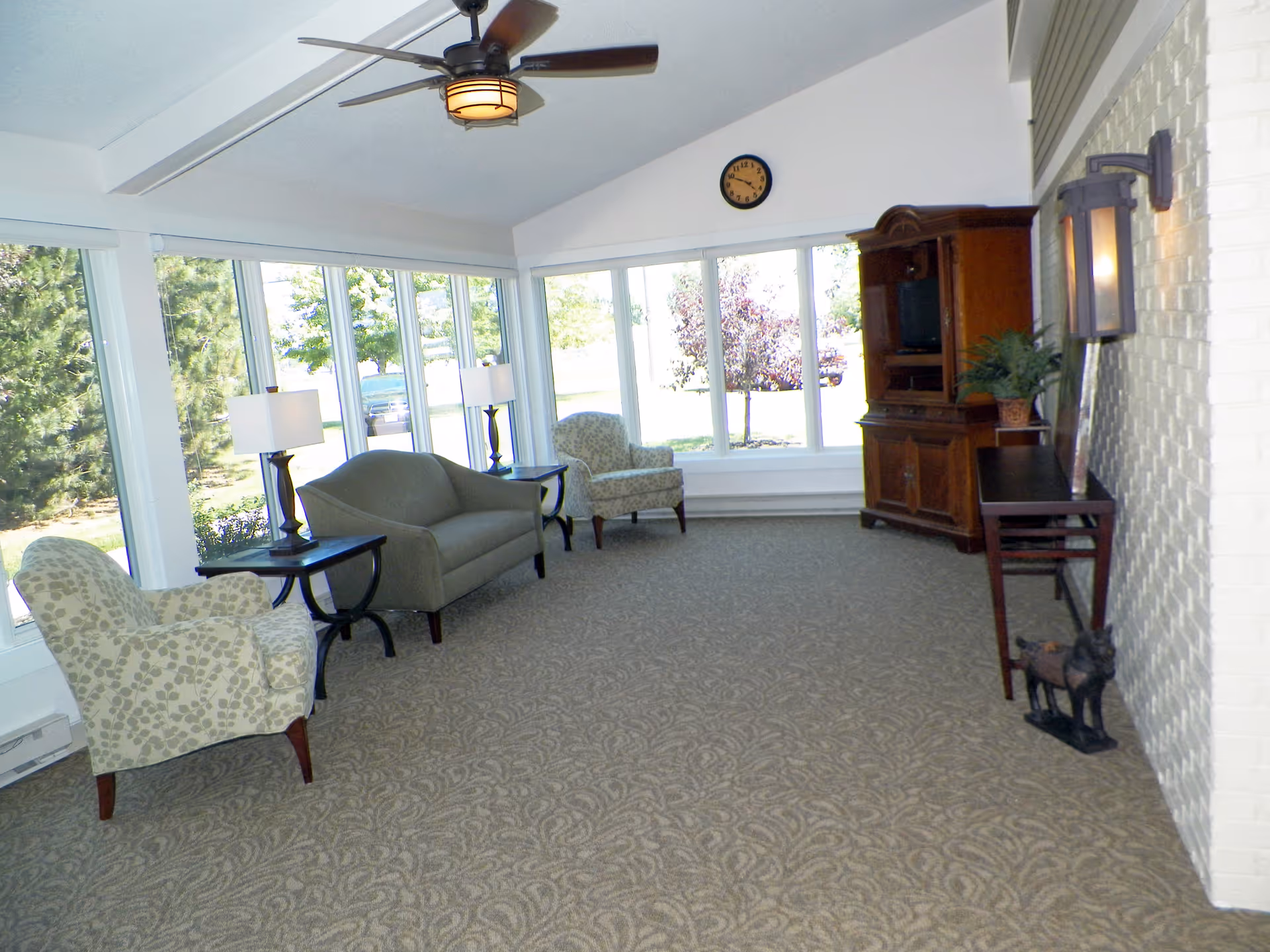 A bright living room with large windows letting in natural light. The room has a patterned carpet, two armchairs with a leaf pattern, a gray loveseat, two side tables with lamps, a wooden entertainment cabinet, a wall clock, a ceiling fan, and a small table with a plant and decorative items near a white brick wall.