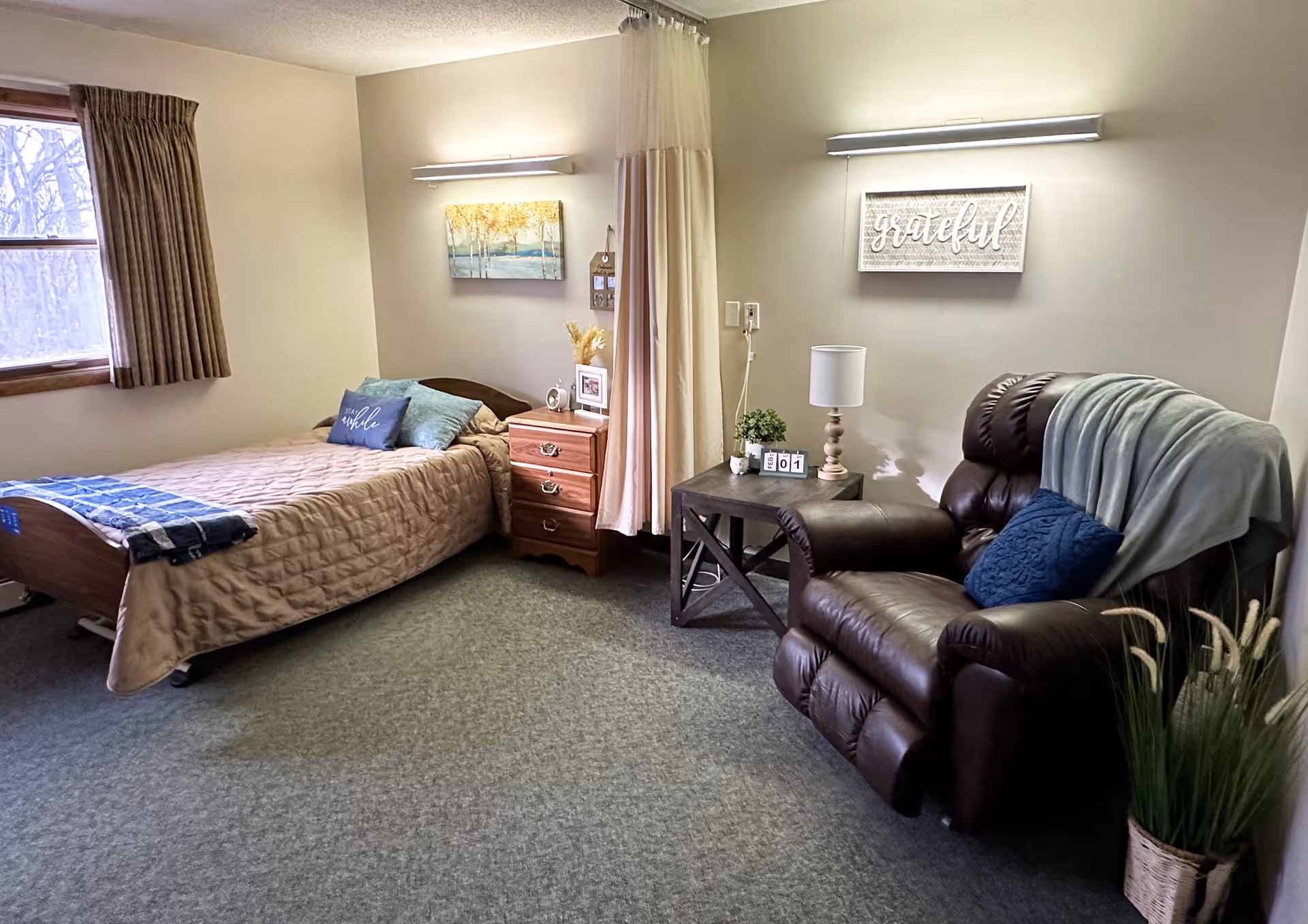 A cozy senior living bedroom with a single bed covered in a beige quilt, a wooden nightstand with a clock and framed photo, a brown leather recliner with a blue pillow and light blue throw blanket, a side table with a lamp and small plant, and wall art including a painting of trees and a sign that says 'grateful'. A window with brown curtains lets in natural light.