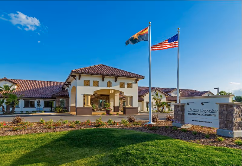 Exterior view of Amber Creek Inn Memory Care Community building with a covered entrance, two flagpoles displaying the Arizona state flag and the American flag, landscaped greenery, and a sign with the facility's name.