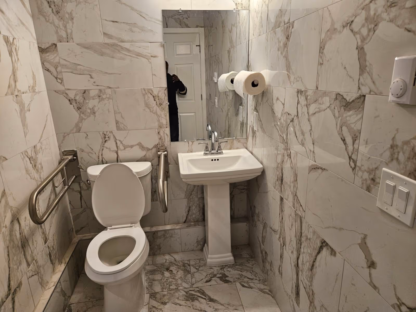 Small marble-tiled bathroom with a toilet (lid up) flanked by stainless steel grab bars, a pedestal sink under a mirror, and two toilet paper rolls on the wall.