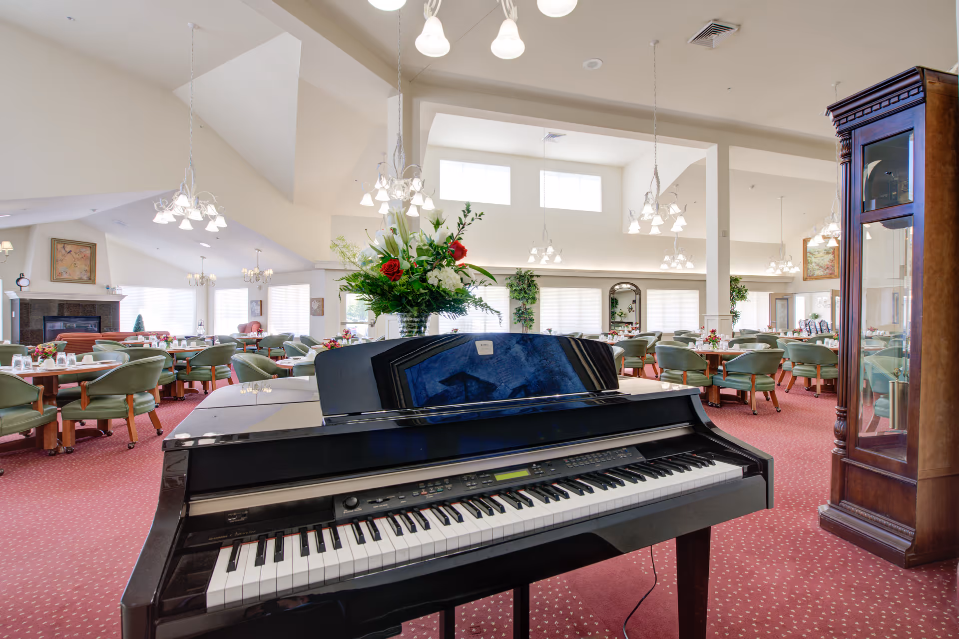 Interior dining room with a piano in the foreground, round dining tables and green chairs under chandeliers.