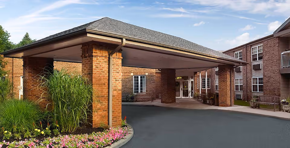 Covered brick entrance to senior living facility with drive, benches, and landscaping.