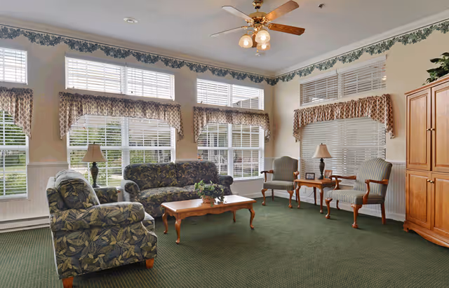 Communal living room with patterned sofas and armchairs, a wooden coffee table, side tables with lamps, and large windows with blinds and valances.