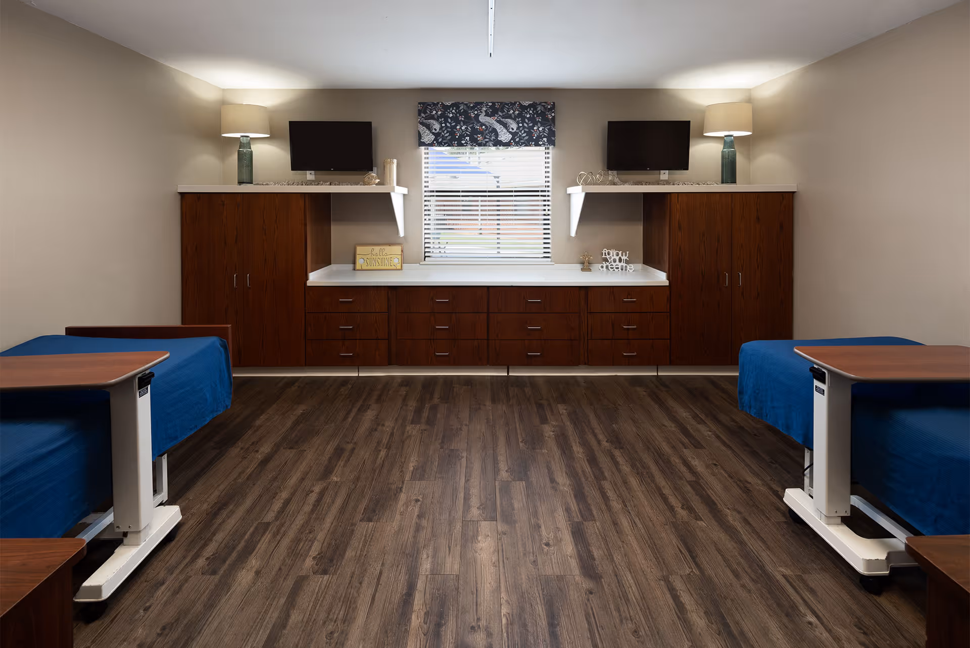 A symmetrical room with two hospital-style beds on either side, each covered with a blue blanket and accompanied by an adjustable overbed table. The back wall features wooden cabinets and drawers with white countertops, two table lamps with green bases and beige shades, two small wall-mounted televisions, and a window with blinds and a patterned valance in the center.