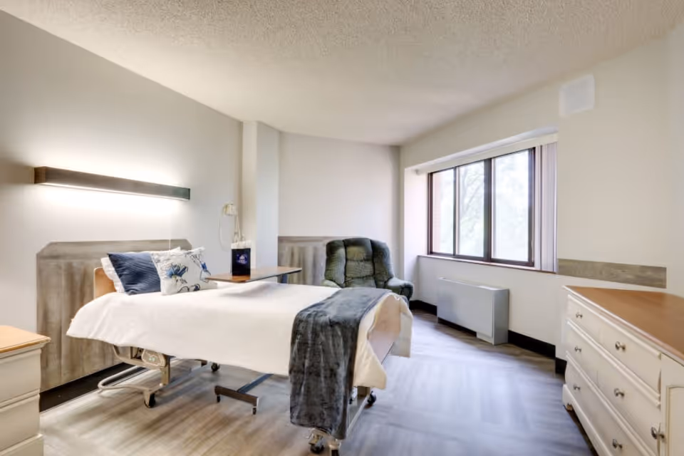 A bright and clean bedroom in a senior living facility featuring a hospital-style bed with white bedding and blue accent pillows, a green upholstered armchair near a large window with vertical blinds, a wooden dresser, and a bedside table with a mounted light fixture above the bed.