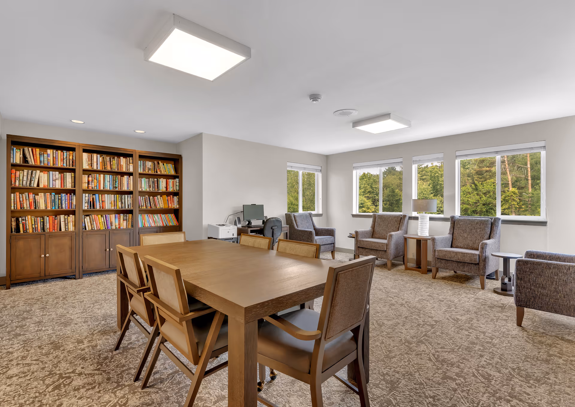 Well-lit communal lounge with a large wooden table and chairs, a bookshelf against one wall, and upholstered armchairs by windows overlooking trees.