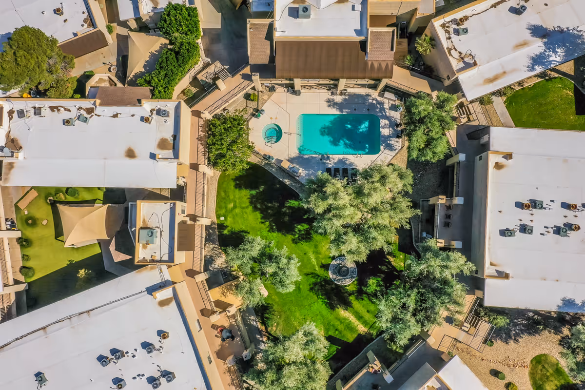 Aerial view of Scottsdale Village Square showing a central courtyard with a swimming pool, hot tub, green lawn, trees, and surrounding buildings with white roofs.
