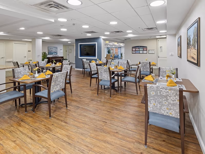 A dining room in a senior living facility with multiple wooden tables and chairs arranged neatly. Each table is set with yellow folded napkins, glasses, and small flower vases. The room has wood flooring, white walls adorned with framed pictures, and a ceiling with recessed lighting. A large flat-screen TV is mounted on a blue accent wall in the background.