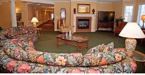A cozy living room area in a senior living facility featuring floral patterned sofas arranged around a wooden coffee table. The room includes a fireplace with decorative items on the mantel, a flat-screen TV on a wooden stand, a grandfather clock, and several lamps providing warm lighting. Large windows with blinds allow natural light into the space.