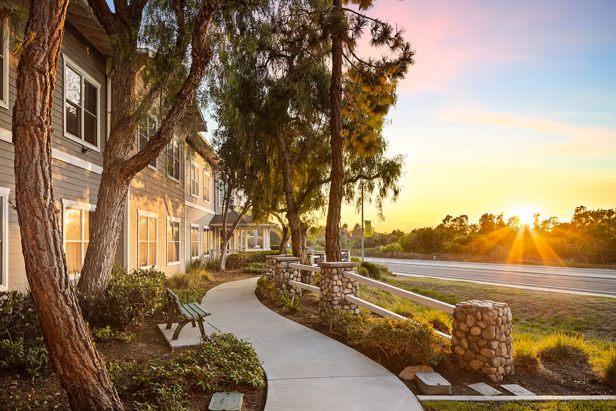 A paved walkway curves alongside a building with multiple windows, bordered by trees, shrubs, and a stone fence. The sun is setting in the background, casting a warm golden light over the scene.