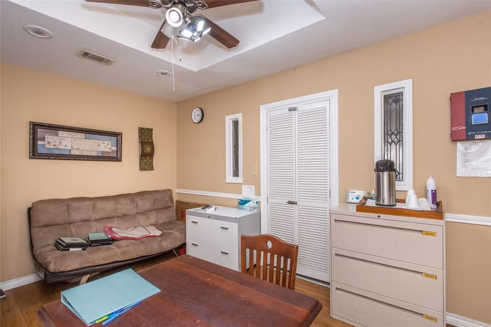 Small communal room with a couch, wooden table and chairs, filing cabinets, and a coffee urn against beige walls.
