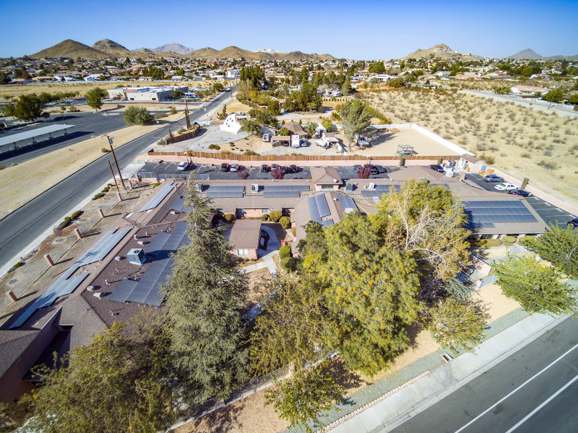 Aerial view of Valley Crest Memory Care facility showing multiple single-story buildings with solar panels on the roofs, surrounded by trees and desert landscape. The facility is located near a road with parked cars and open desert areas with mountains in the background under a clear blue sky.