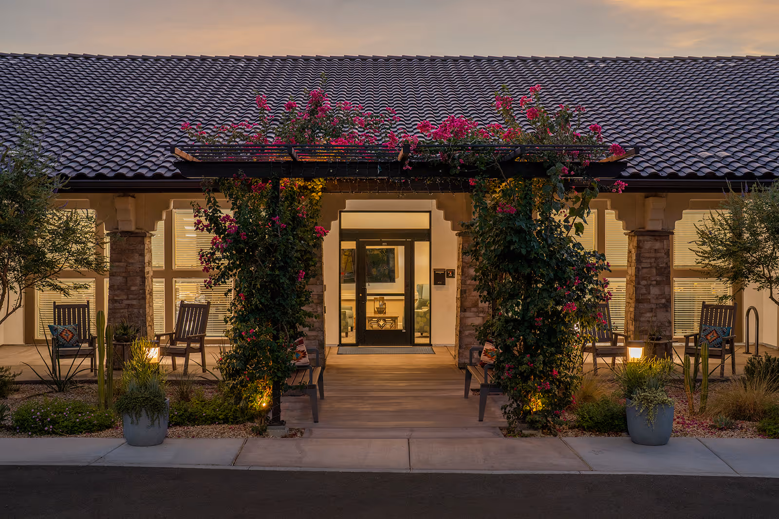 Front entrance of a memory care building at dusk with a vine-covered pergola, warm exterior lighting, potted plants, and outdoor seating.