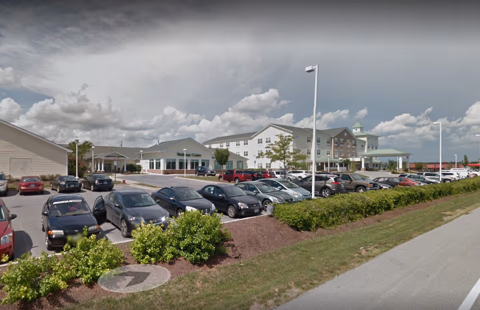 Exterior view of Lorien Taneytown facility showing a large parking lot filled with cars, landscaped bushes, and a multi-story building with a covered entrance and a sign for the Wellness Center. The sky is partly cloudy.