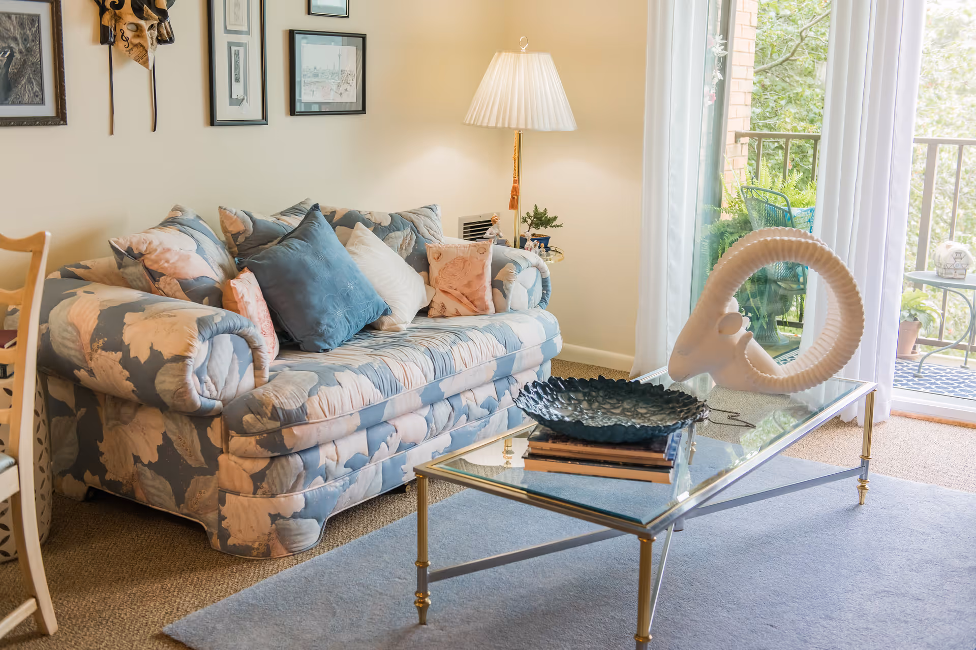 A cozy living room with a floral patterned sofa adorned with multiple cushions in blue, white, and pink. A glass coffee table with gold legs holds a decorative blue bowl and a white ram's head sculpture. A floor lamp with a pleated shade stands beside the sofa. The room has framed artwork on the wall and a sliding glass door leading to a balcony with outdoor furniture and plants.
