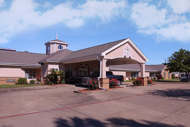 Exterior view of a single-story senior living facility building with a covered entrance driveway. The building has beige walls, a brown shingled roof, and a small cupola on top. There are landscaped bushes and flowers near the entrance, and a pickup truck is parked under the covered area. The sky is partly cloudy.