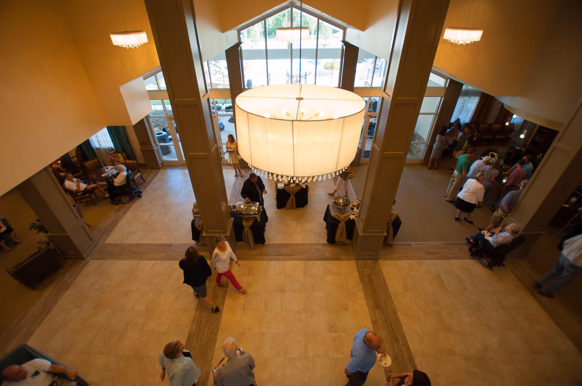 View from above of a spacious senior living facility lobby with a large chandelier hanging from the ceiling. Several people are gathered around tables with food and drinks, while others are seated or walking through the area. Large windows and glass doors provide natural light and a view of the outdoor area.