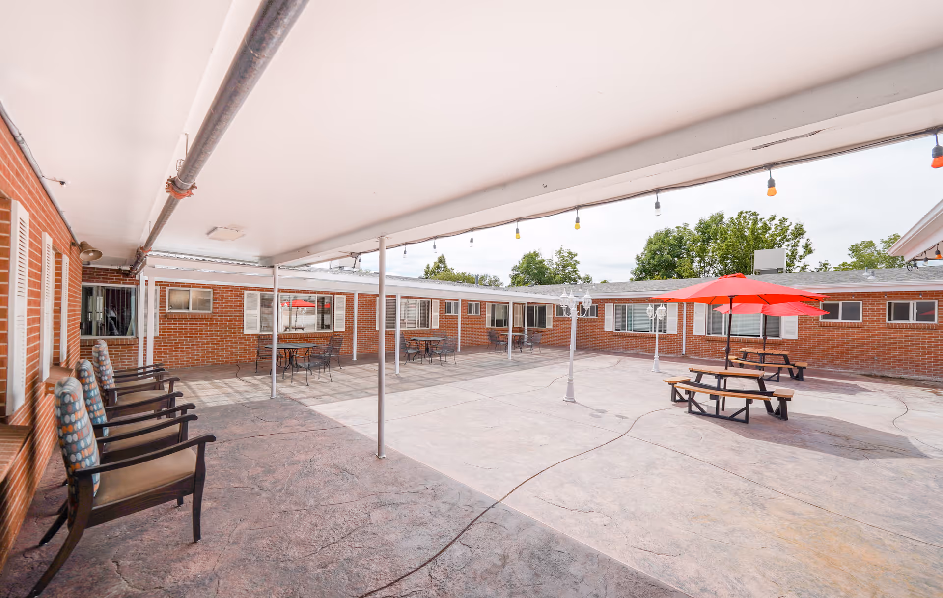 Outdoor courtyard area of a nursing and rehabilitation center with brick walls surrounding it. There are several chairs lined up under a covered patio on the left side, and picnic tables with red umbrellas in the open area. String lights hang from the ceiling of the covered patio and there are trees visible in the background.