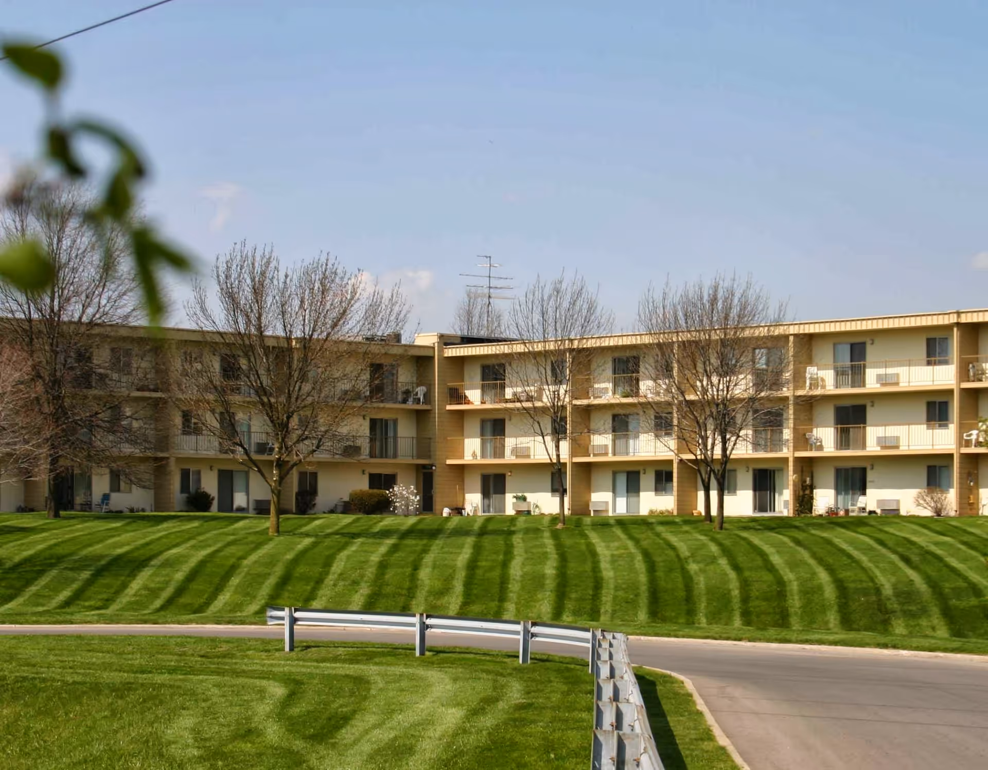 Exterior view of a three-story senior living facility building with balconies, surrounded by neatly mowed green grass and leafless trees under a clear blue sky.