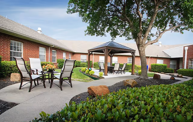 Outdoor courtyard area of a senior living facility with a concrete pathway, patio chairs and tables, a wooden gazebo, green bushes, a large tree, and red brick buildings in the background under a clear blue sky.
