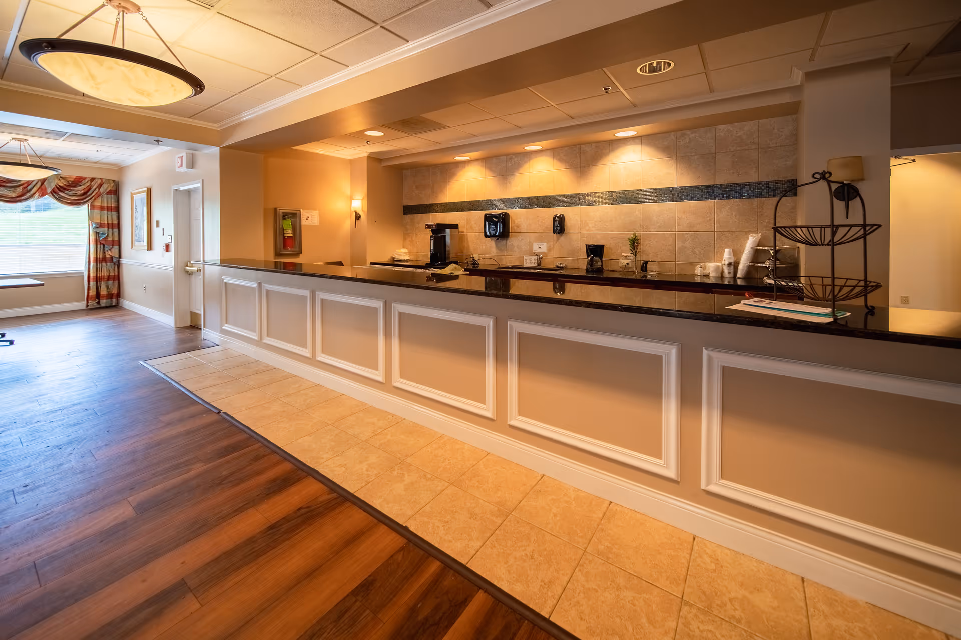 A well-lit reception desk and lobby area with a long counter, tiled backsplash and a coffee station, open to a windowed seating area.