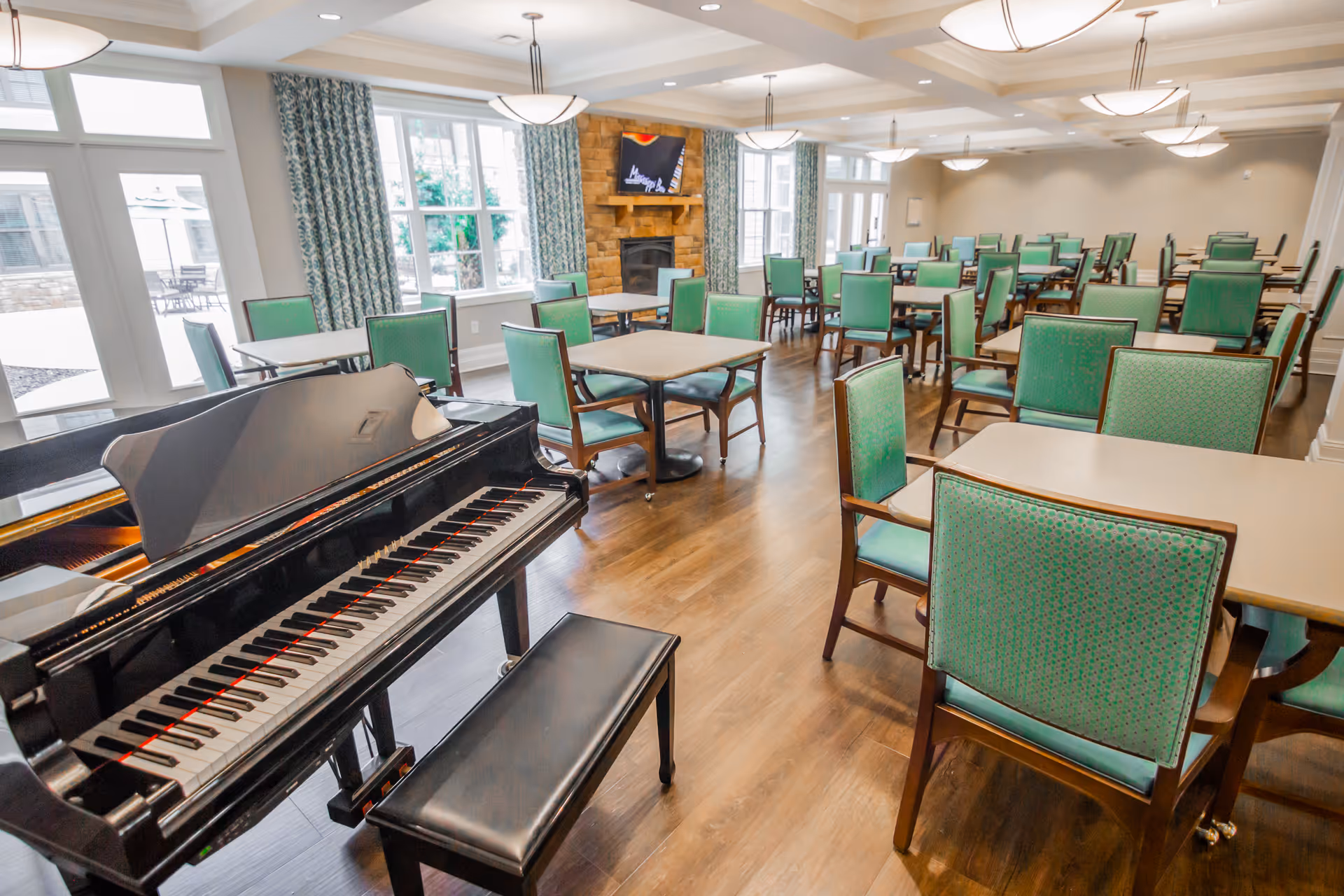 Bright communal dining/activity room with a grand piano in the foreground, multiple tables and green-upholstered chairs, windows and a fireplace.
