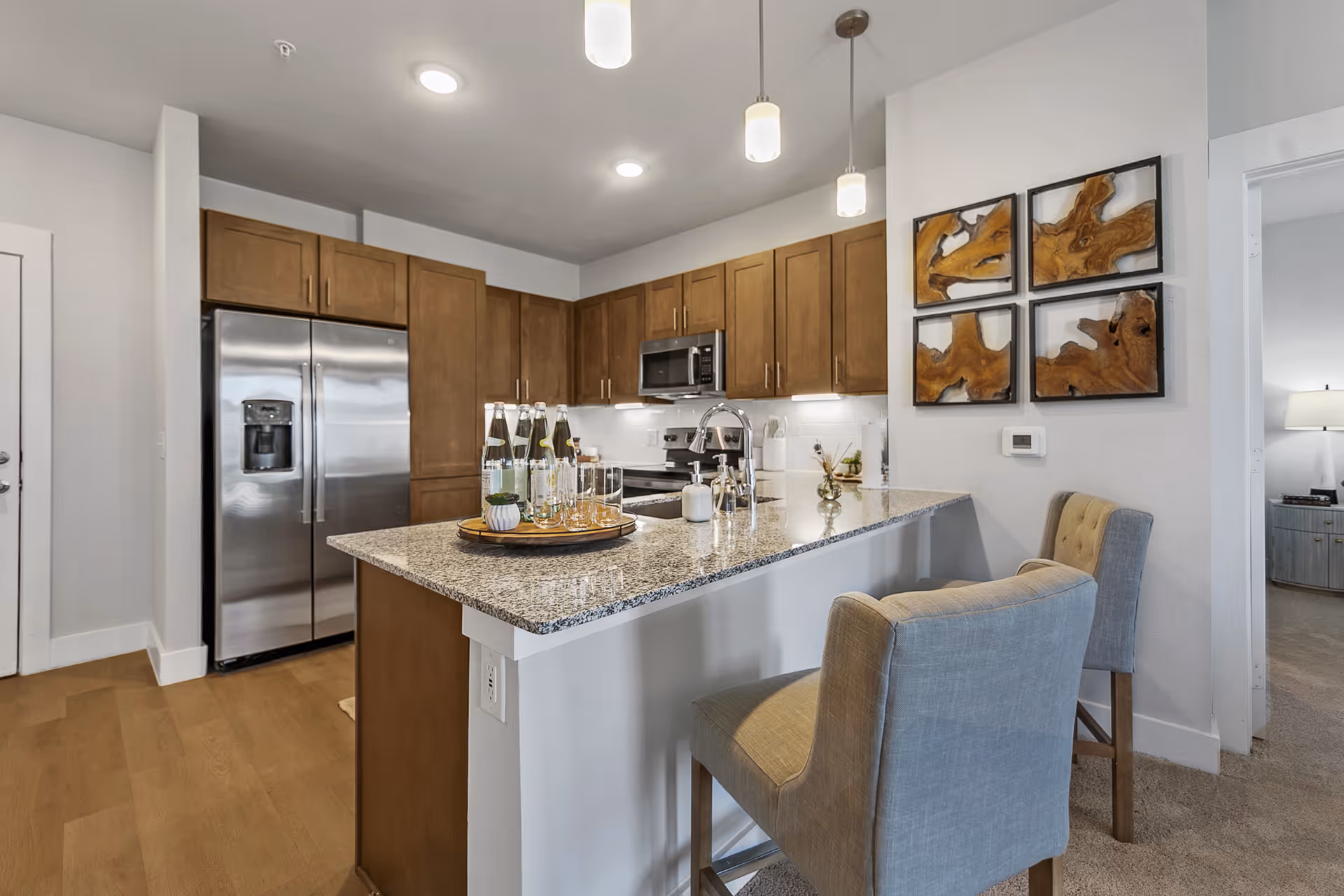 Modern open kitchen with a granite-topped island, stainless steel refrigerator, wooden cabinets, and two upholstered bar chairs.