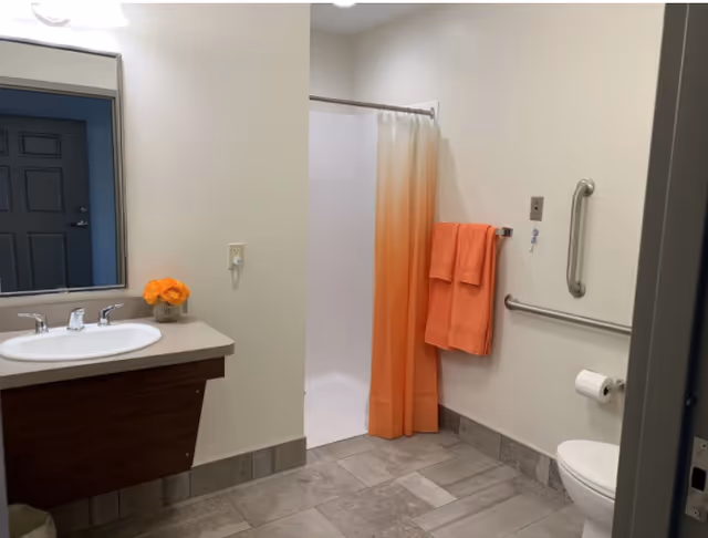 A clean bathroom with a white sink and countertop on the left, a mirror above the sink, and a small vase with orange flowers. In the background, there is a shower with a white and orange gradient shower curtain. On the right wall, there are two orange towels hanging on a towel rack, a grab bar, and a toilet with a toilet paper holder. The floor is tiled in gray tones.