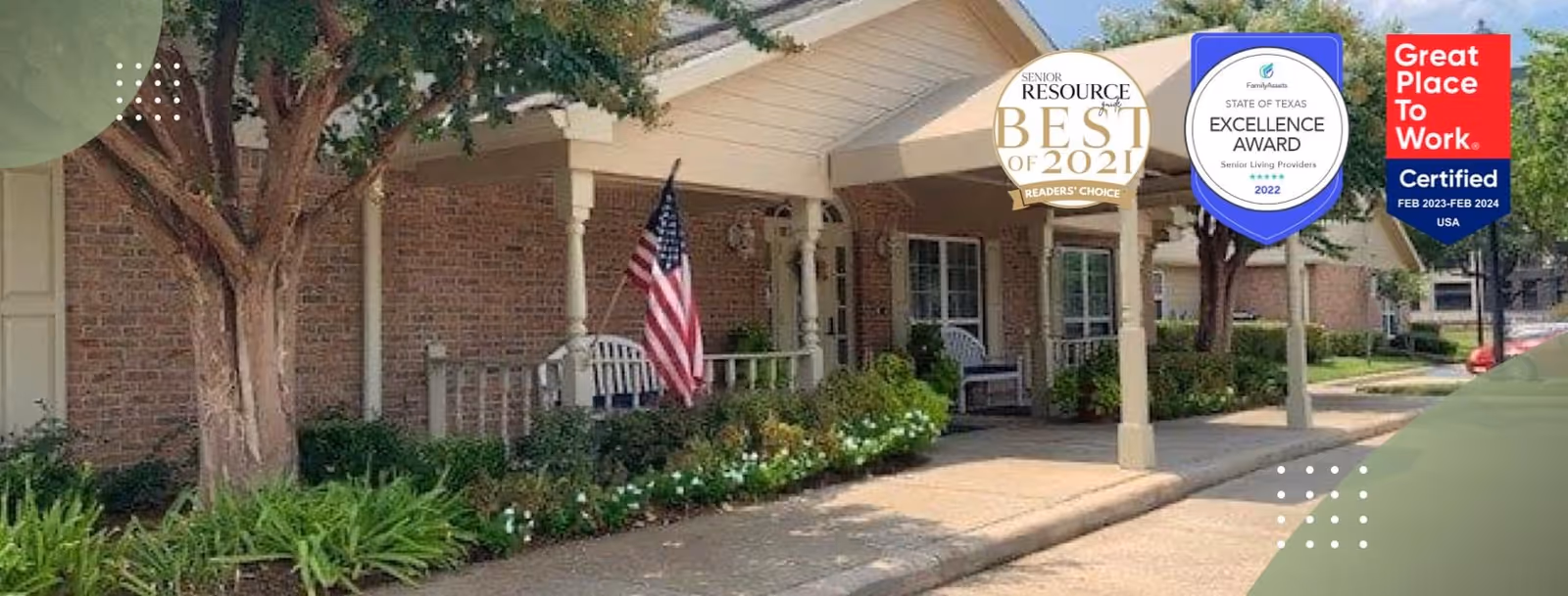 Exterior view of a senior living facility with a covered entrance, brick walls, and a porch area with chairs. There is an American flag displayed near the entrance, surrounded by green shrubs and flowers. Trees and a sidewalk are visible along the driveway.