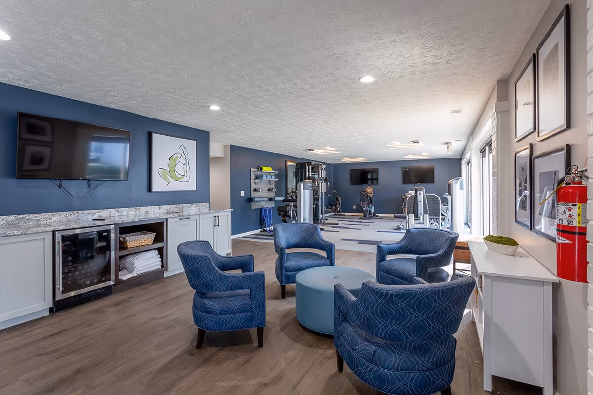 A spacious room with a seating area consisting of four blue patterned armchairs arranged around a round blue ottoman. The room has wood flooring and a blue accent wall with a mounted flat-screen TV and framed artwork. In the background, there is exercise equipment including stationary bikes and weight machines. A countertop with cabinets and a small refrigerator is visible on the left side, and a white console table with framed pictures and a fire extinguisher is on the right side near large windows.