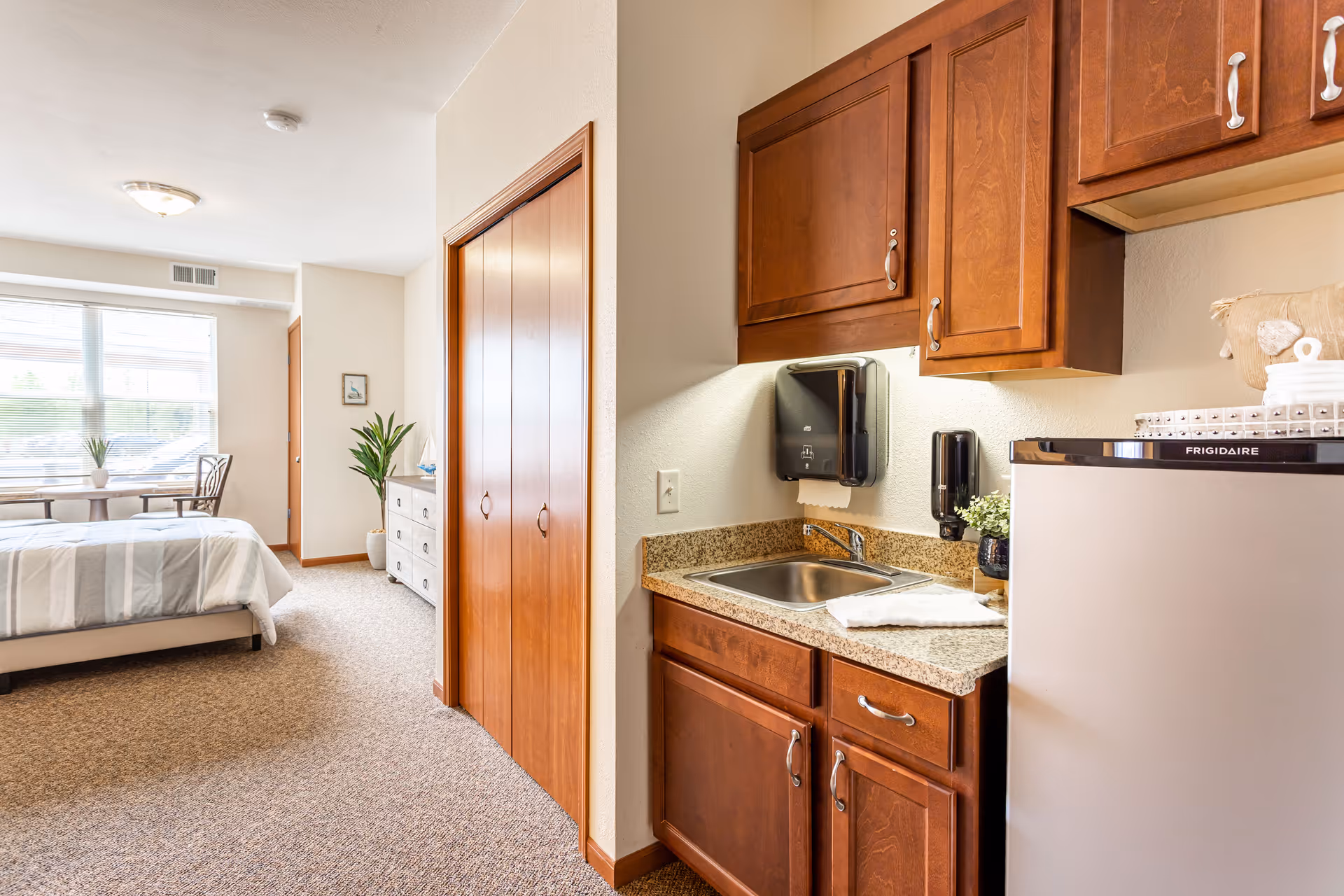 Interior view of a senior living facility room featuring a small kitchenette with wooden cabinets, a sink, and a mini refrigerator on the right side. The room extends to a sleeping area with a bed, a dresser, a small table with two chairs near a large window, and a closet with wooden sliding doors. The space is well-lit with natural light coming through the window.