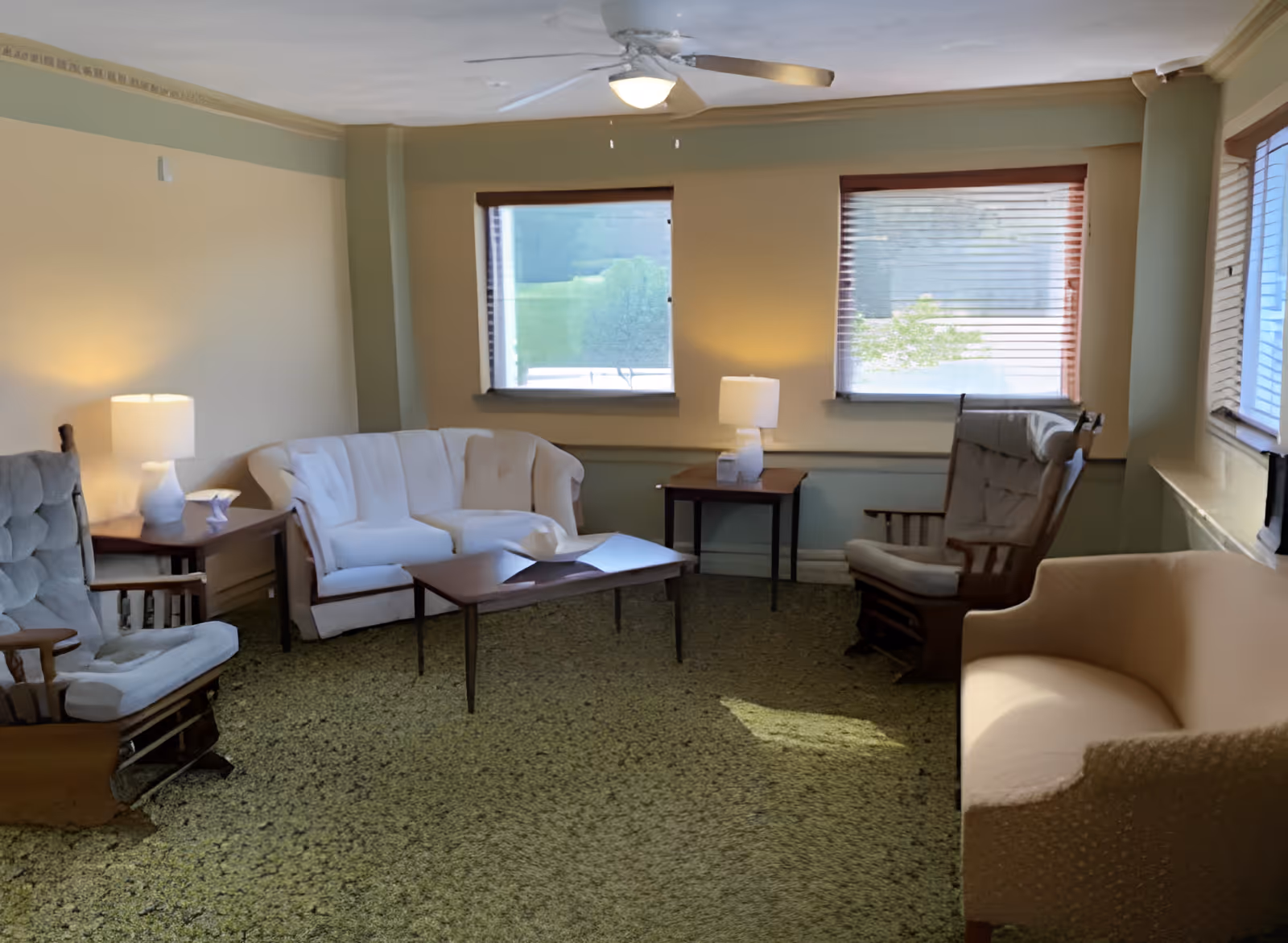 A cozy living room with upholstered sofas and armchairs arranged around coffee tables, lamps, and windows with blinds.