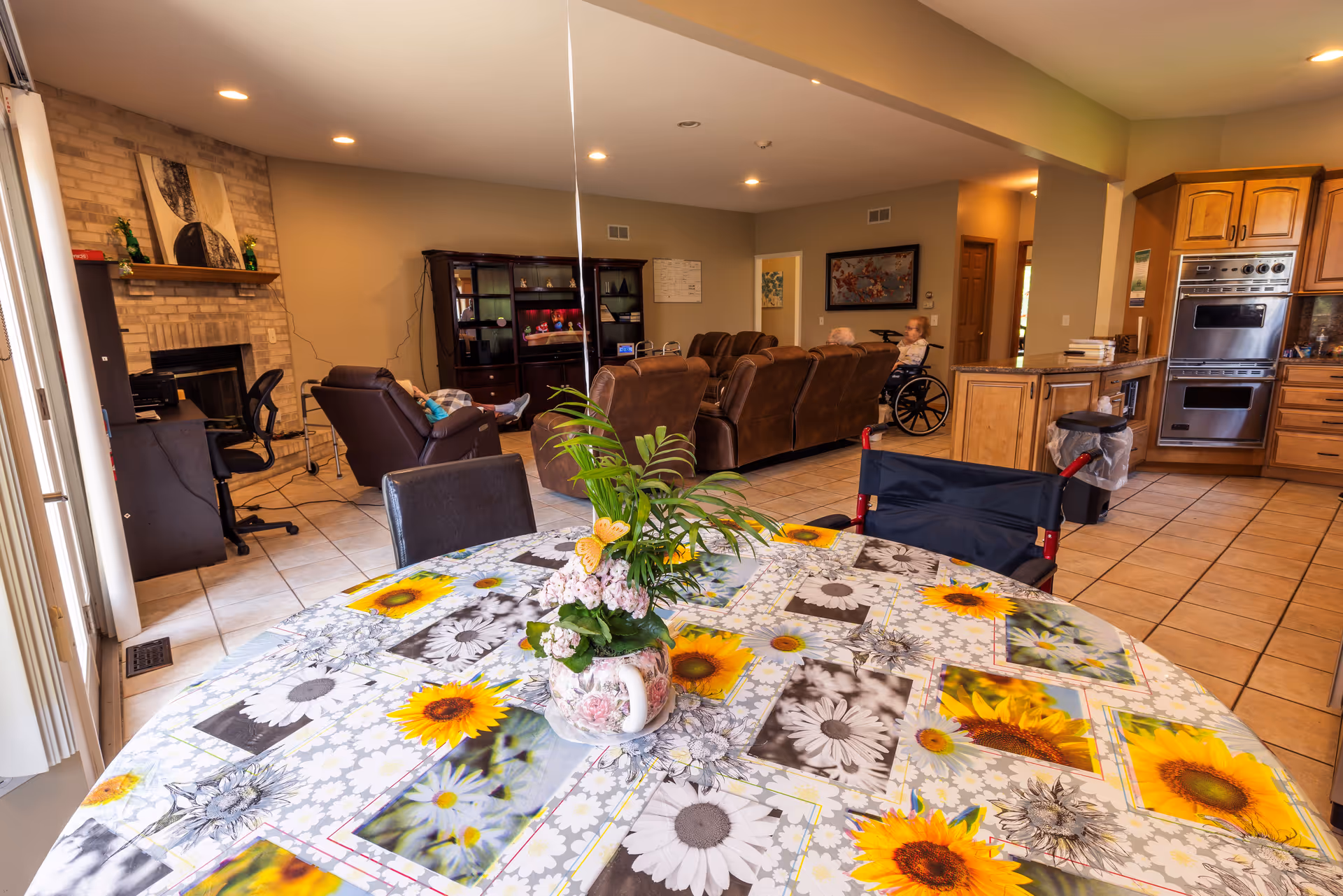 A bright and spacious common area in a senior living facility featuring a round table with a floral tablecloth and a vase of flowers in the foreground. In the background, there are brown leather recliners and sofas, a TV cabinet, and two elderly individuals, one seated in a wheelchair. The room also includes a kitchen area with wooden cabinets and stainless steel appliances, tiled flooring, and a brick fireplace on the left side.