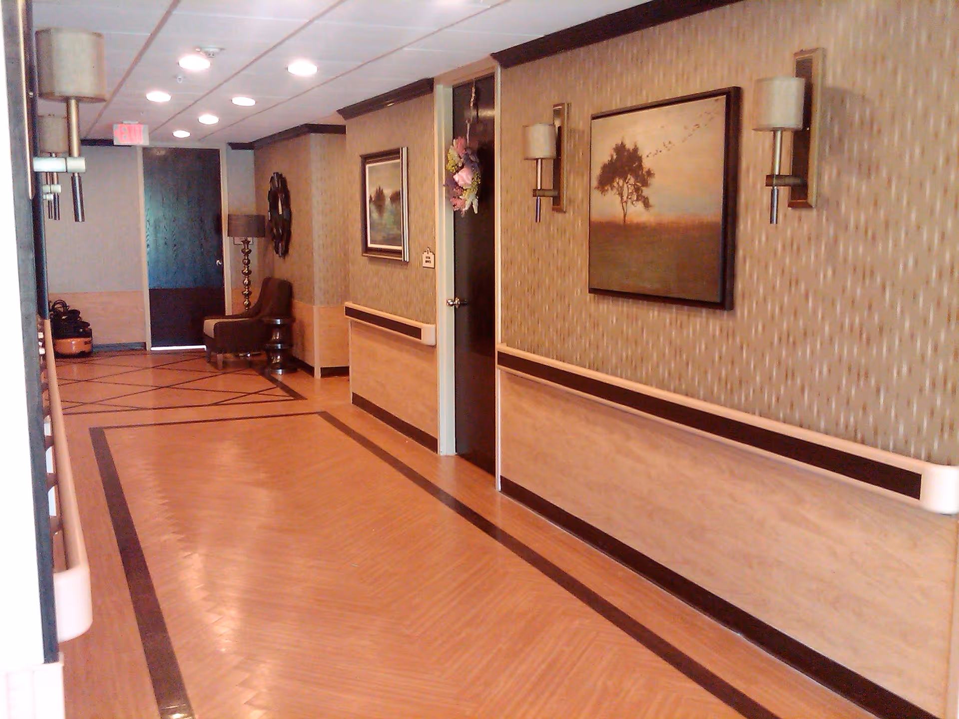 A well-lit hallway in a senior living facility with wood-patterned flooring and beige patterned wallpaper. The walls have wooden paneling with handrails. There are framed landscape paintings and wall sconces on the right wall. At the end of the hallway, there is a brown chair, a floor lamp, and a dark door.