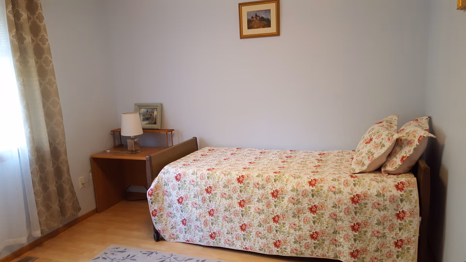 A small bedroom with a single bed covered in a floral bedspread, a bedside table with a lamp and framed pictures, and a window with patterned curtains.