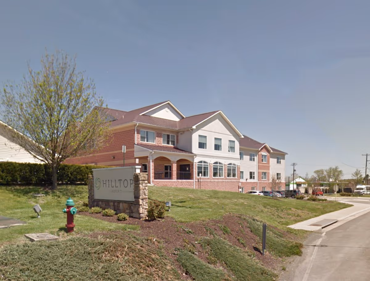 Exterior view of a two-story senior living facility named Hilltop, with a brick and white facade, a red roof, a landscaped grassy area with a tree and a fire hydrant in front, and a parking lot to the right under a clear blue sky.