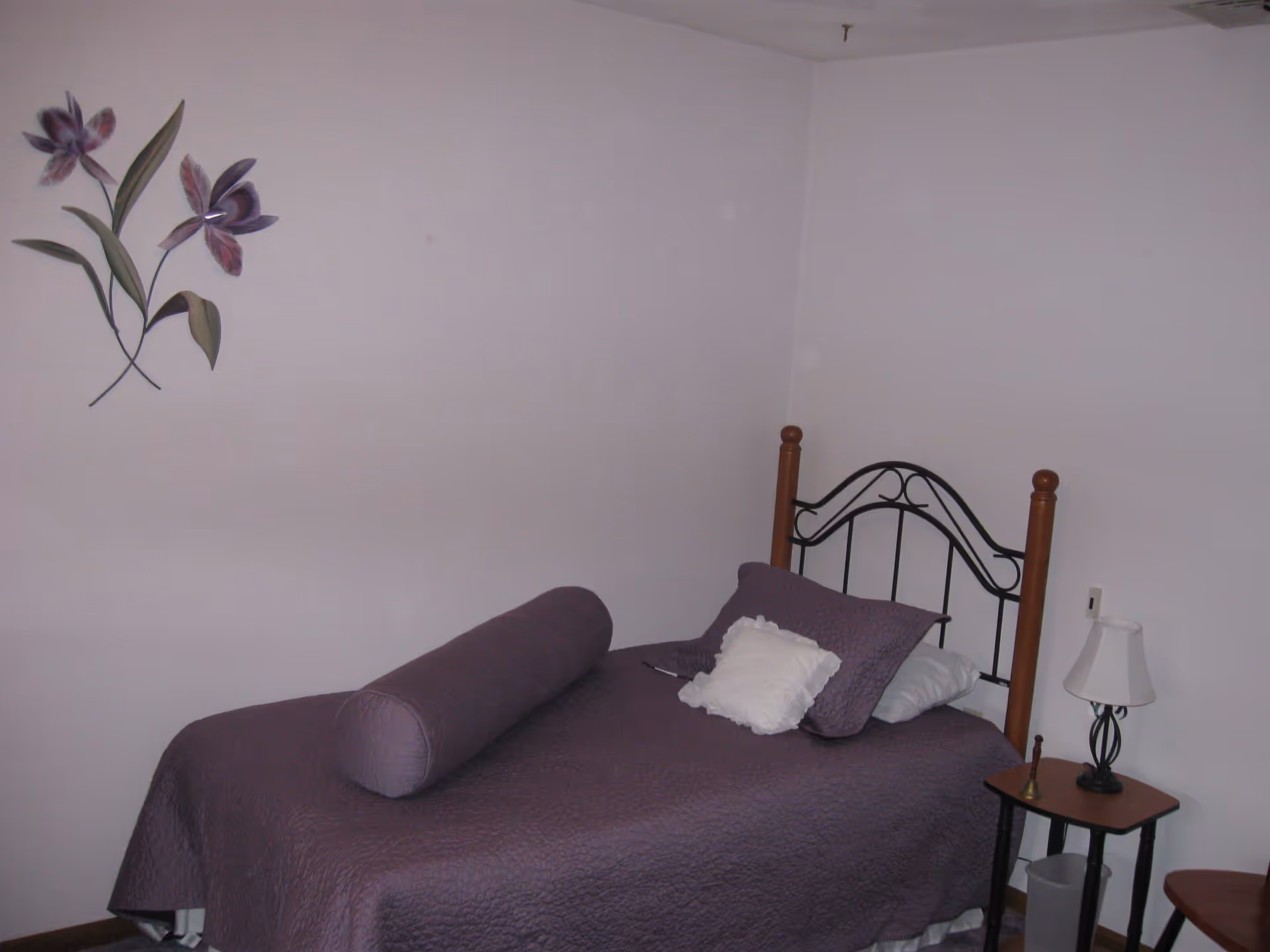 A small bedroom with a single bed covered in a purple quilt and matching pillows. There is a decorative metal and wood headboard. A small wooden side table next to the bed holds a lamp and a small bell. The walls are plain white with a metal wall decoration of purple flowers.